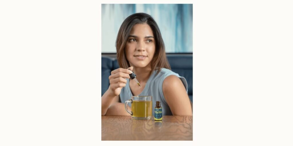 A young woman adds a dose of KeySlim Drops into a glass mug using a dropper. A small bottle of the supplement sits on the wooden table in front of her. The setting is bright and modern, emphasizing a simple, healthy daily routine.